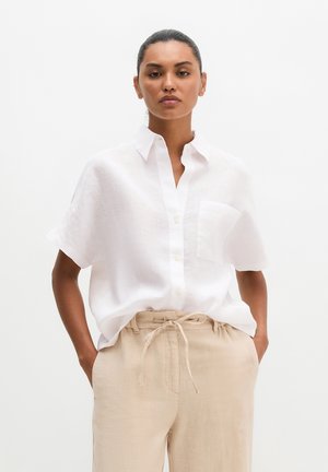 Femme portant une chemise blanche à manches courtes boutonnée, rentrée dans un pantalon beige à cordon, debout les mains dans les poches.