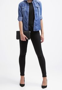 Camisa de mezclilla con mangas enrolladas, top negro ajustado, jeans skinny negros, clutch negro y tacones de aguja. Diseño simple y elegante.