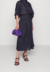 Nainen, joka pukeutunut tummansiniseen napitettuun paitapuseroon ja hameeseen, pitää violetista käsilaukusta kiinni ja käyttää mustia korkokenkiä yksinkertaista taustaa vasten.