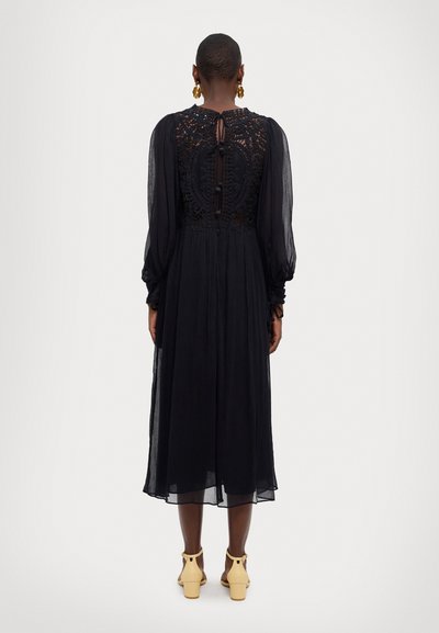 Black midi dress with long, sheer puff sleeves. Lace detailing at the back, keyhole opening, and flowing fabric. Pairing with yellow heels.