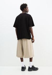 Černé oversized tričko, béžové široké šortky, černé tenisky a bílé kotníkové ponožky. Pozadí je světlé a minimalistické.