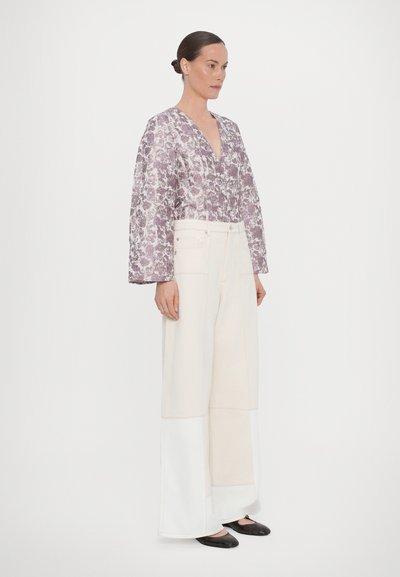 Paarse bloemenshirt met volumineuze mouwen, gecombineerd met brede crèmekleurige broeken met kleurblokontwerp en zwarte flats.