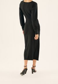 Svart långärmad klänning med en åtsittande silhuett och sidofållningar, tillverkad av mjukt tyg, knälång, matchad med svarta öppna klackskor.