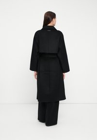 Woman with long brown hair wearing a long black coat with wide sleeves, standing facing away against a plain white background.