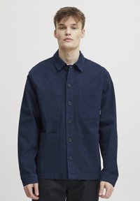 Navy blue, button-up jacket made of cotton, featuring two front pockets, a collar, and long sleeves with a clean finish.