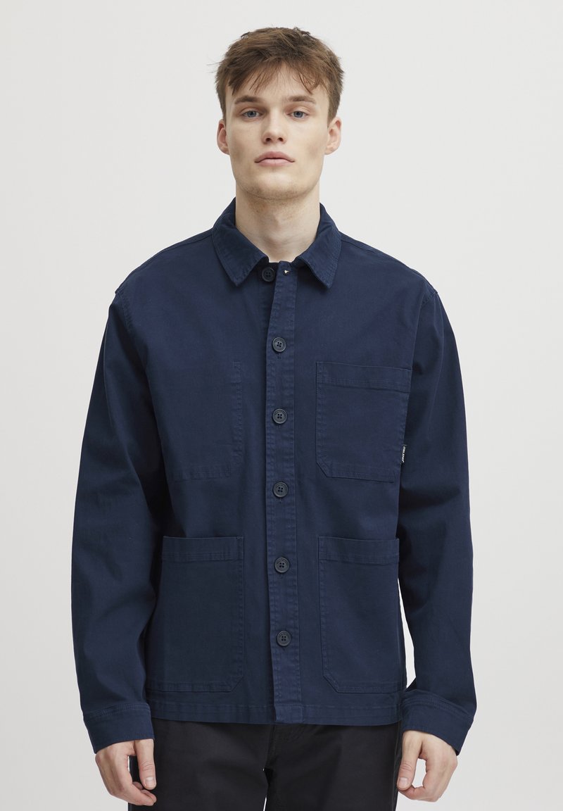 Navy blue, button-up jacket made of cotton, featuring two front pockets, a collar, and long sleeves with a clean finish.