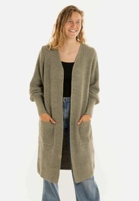 Vrouw glimlachend, gekleed in een lange grijze gebreide cardigan over een zwarte top en blauwe jeans, met handen in de zakken van de cardigan.