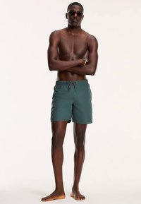 Pantaloni scurți de baie verzi, realizați dintr-un material ușor, cu talie elastică, șnur de strângere și buzunare laterale. Modelul este accesorizat cu ochelari de soare.