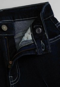 Ciemnoniebieskie dżinsy z zamkiem błyskawicznym i zapięciem na guzik. Wnętrze wykończone jasnoniebieską tkaniną oraz kontrastującymi przeszyciami.