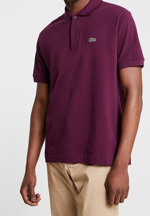 Homme portant un polo à manches courtes de couleur violet foncé avec un petit logo de crocodile vert sur la poitrine gauche et un pantalon beige.
