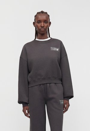 Femme aux cheveux tressés portant un sweat-shirt gris foncé New Balance Athletics et un pantalon de survêtement assorti sur un fond uni.