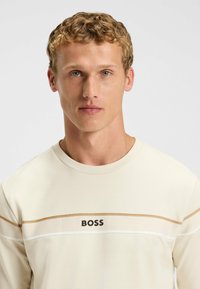 Beige langemouwen shirt met een ronde hals, twee horizontale strepen in lichtbruin en wit, met het woord "BOSS" in zwarte print.
