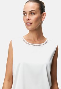 White sleeveless top with a round neckline featuring a delicate cut-out pattern along the edge. Smooth, soft fabric with a relaxed fit.