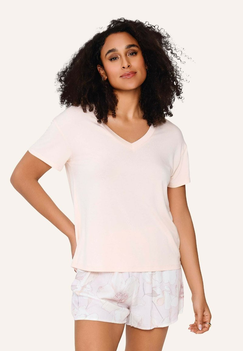 Femme aux cheveux bouclés portant un haut rose clair à col en V et manches courtes ainsi qu'un short blanc à motifs floraux, debout avec une main sur la hanche.