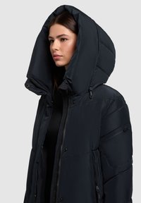 Zwarte pufferjas met een grote, oversized capuchon, ritssluiting en drukknoopsluitingen, zijzakken en een gladde, matte textuur.