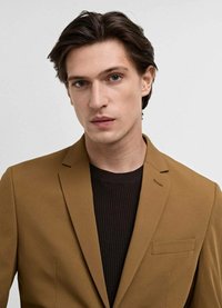 Brown blazer with a slim fit, single-breasted design, notch lapels, and a pocket. Worn over a black ribbed sweater. Neutral background.