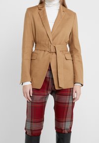 Beżowa marynarka z paskiem, o gładkiej teksturze, dopasowanym kroju i dwóch przednich kieszeniach, w połączeniu z czerwonymi spodniami w kratę.