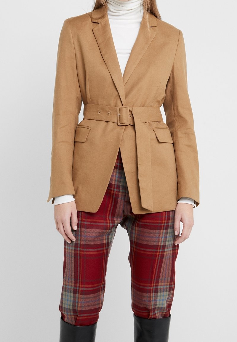Beżowa marynarka z paskiem, o gładkiej teksturze, dopasowanym kroju i dwóch przednich kieszeniach, w połączeniu z czerwonymi spodniami w kratę.