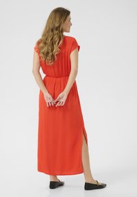 Red maxi dress with short sleeves and a cinched waist, featuring a side slit and smooth fabric. Worn with black flat shoes.