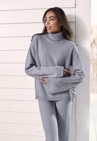 Sivi pleteni pulover s turtleneck ovratnikom in širokimi rokavi, v kombinaciji s prilagojenimi hlačami. Mehak tekstur, sproščen kroj, rebrast rob na ovratniku in manšetah.