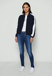 Casaco bombástico azul-marinho com riscas brancas nas laterais, fecho de zip, e punhos canelados, combinado com uma t-shirt branca e jeans skinny azuis. Ténis brancos.