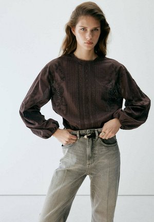 Joven mujer con cabello largo y castaño, que lleva una blusa oscura bordada y unos vaqueros de tiro alto de color gris claro con un cinturón negro, de pie con las manos en las caderas.