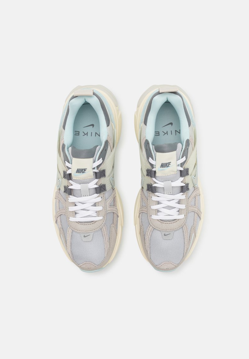 Un par de zapatillas Nike de color gris, beige y azul claro con cordones blancos y suelas gruesas, vistas desde arriba sobre un fondo blanco.