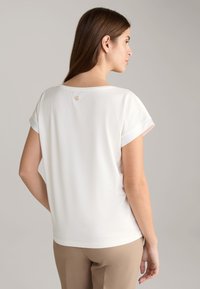 White short-sleeve top with a round neckline, featuring a small logo accent on the back. Smooth texture, relaxed fit.
