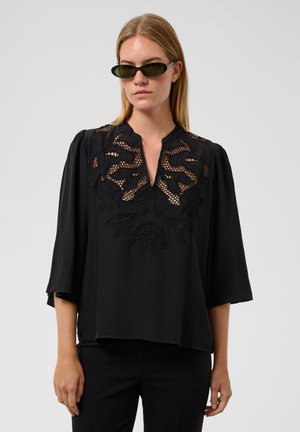 Femme portant des lunettes de soleil foncées et un chemisier noir avec une dentelle élaborée et une broderie florale sur le haut de la poitrine et les épaules.