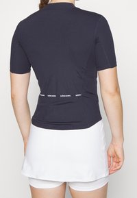 Top atlético de manga corta en azul marino con textura acanalada, presenta un dobladillo curvado y una banda de logo blanca en la parte posterior. Se lleva con una falda blanca.