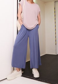 Maglietta rosa chiaro a maniche corte abbinata a pantaloni larghi blu chiaro. La modella indossa sneakers bianche e porta una borsa intrecciata. Outfit semplice e casual.