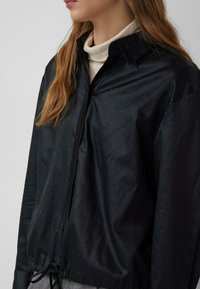 Kobieta ubrana w czarną koszulę z guzikami ozdobioną małymi błyszczącymi kropkami, na kremowym golfie, z widocznymi długimi, jasnobrązowymi włosami.