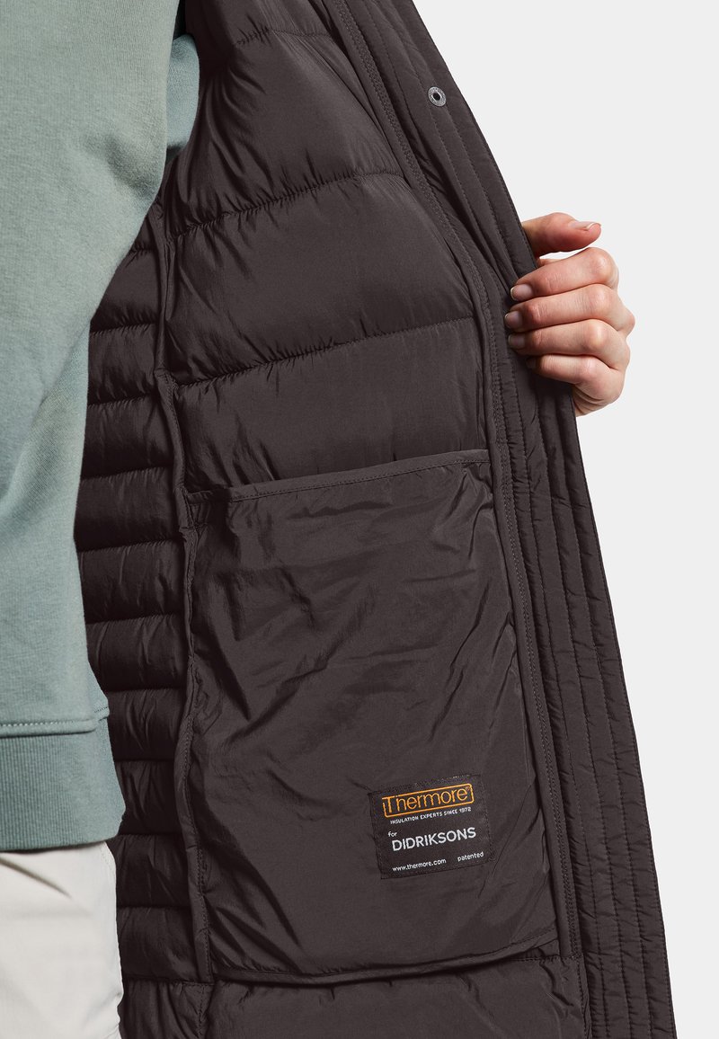 Eine Hand hält eine offene schwarze gesteppte Jacke und zeigt die Innentasche mit dem Thermore-Dämmmaterial-Label.