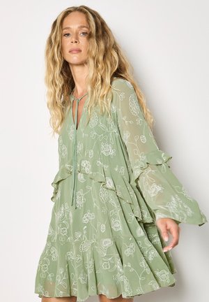 Frau mit langen blonden Haaren, die ein fließendes, salbeigrünes Blumenkleid mit Rüschenschichten und transparenten Glockenärmeln trägt und vor einem schlichten Hintergrund steht.