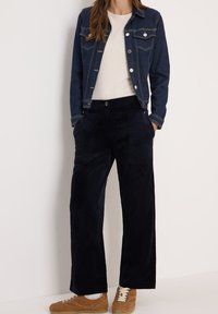 Dunkelblaue Denimjacke mit zwei Brusttaschen, weißem T-Shirt und navyfarbener Cordhose im weiten Schnitt. Beigefarbene Schnürsneaker runden das Outfit ab.