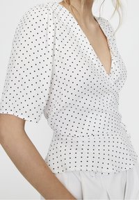 Donna che indossa una blusa avvolgente bianca a maniche corte con pois neri, abbinata a pantaloni bianchi a vita alta, visuale parziale del torso.