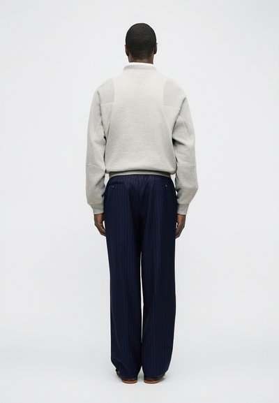 Άνδρας που στέκεται με την πλάτη στην κάμερα, φοράει ανοιχτό γκρι πουλόβερ και σκούρο μπλε παντελόνι με ρίγες και φαρδιά μπατζάκια, πάνω σε ένα απλό ανοιχτό φόντο.