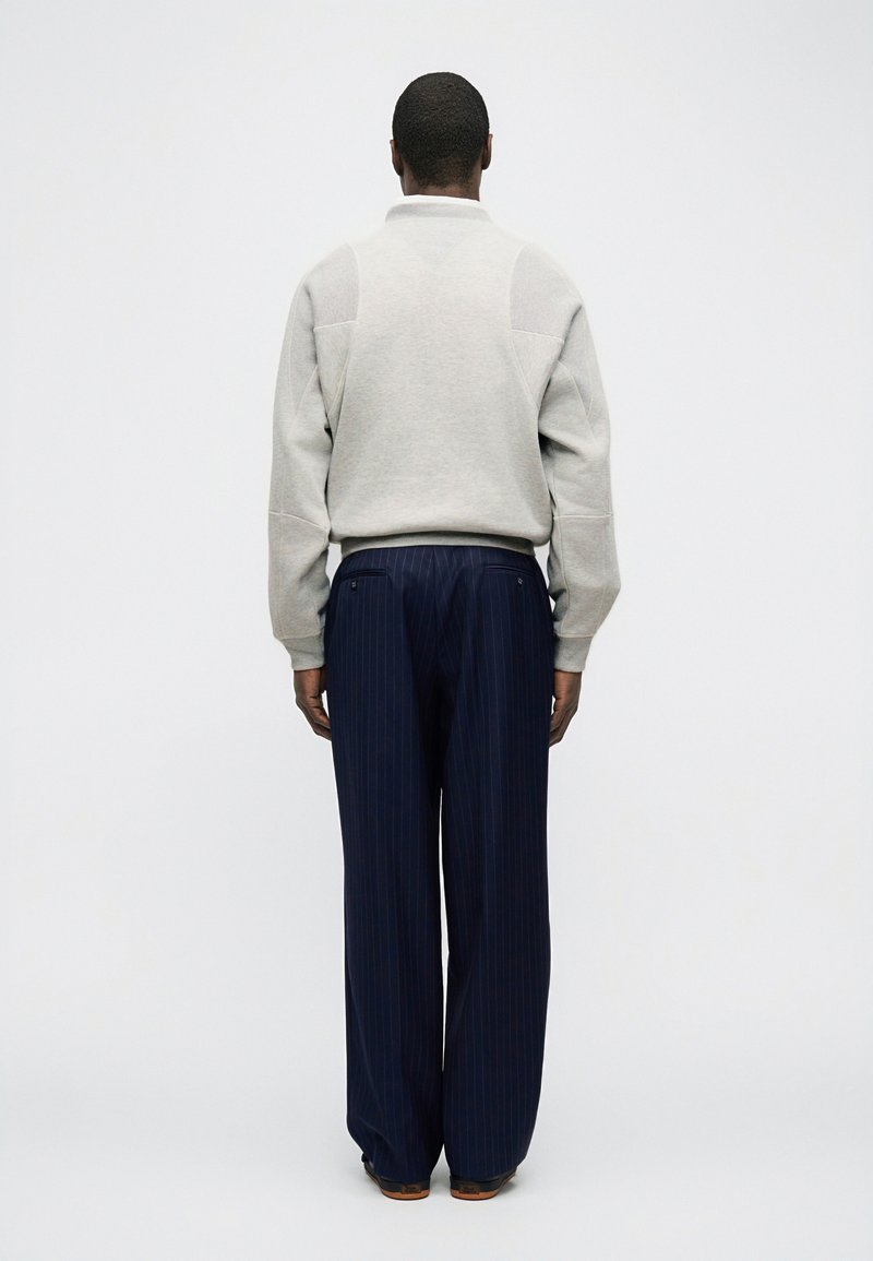 Uomo in piedi di spalle, indossa un maglione grigio chiaro e pantaloni a gamba larga a righe blu scuro su uno sfondo chiaro e uniforme.