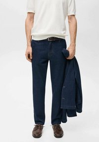 Donkerblauwe denim broek met een rechte pasvorm, gecombineerd met een witte shirt met korte mouwen en bruine schoenen. Jas over de arm gedrapeerd.