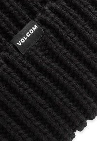 Zwarte gebreide beanie van zacht garen, met een gestructureerd patroon en een zichtbaar wit "Volcom" logo label dat aan de zijkant is gestikt.