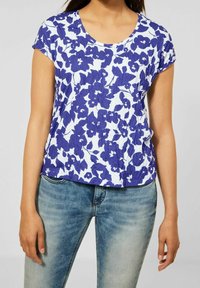 Žena v bílém tričku s krátkým rukávem s modrým květinovým vzorem, které kombinuje se světle modrými vybledlými džíny, na jednobarevném pozadí.
