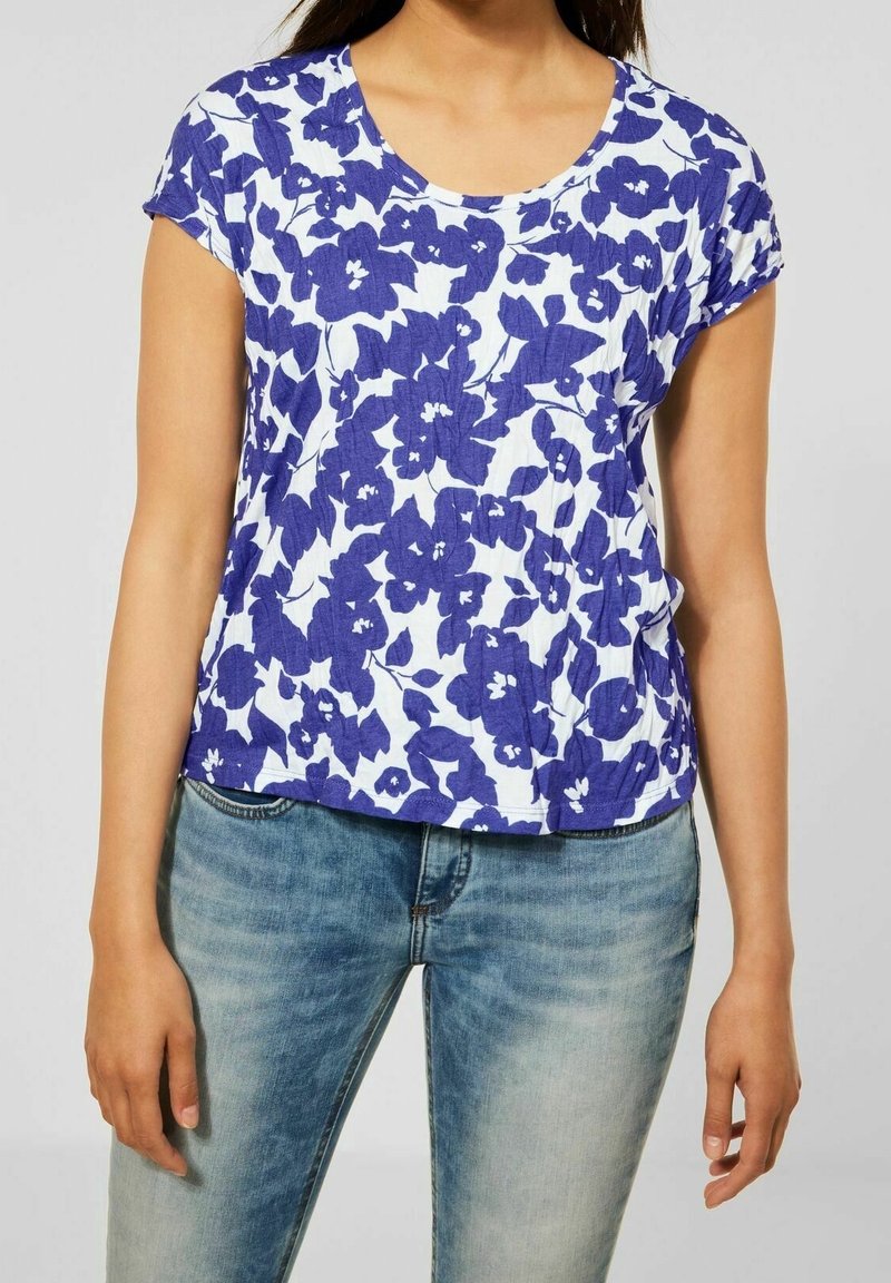 Žena v bílém tričku s krátkým rukávem s modrým květinovým vzorem, které kombinuje se světle modrými vybledlými džíny, na jednobarevném pozadí.
