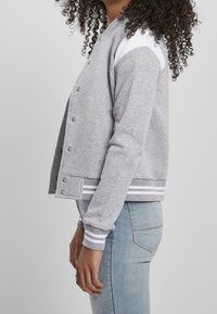 Gray knit varsity jacket with white shoulder accents, snap buttons, and striped ribbed cuffs and hem, shown on a person from the side.