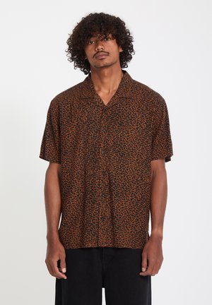 Hombre con cabello rizado que lleva una camisa de manga corta con botones y patrón marrón y negro, y pantalones negros, posando contra un fondo blanco.