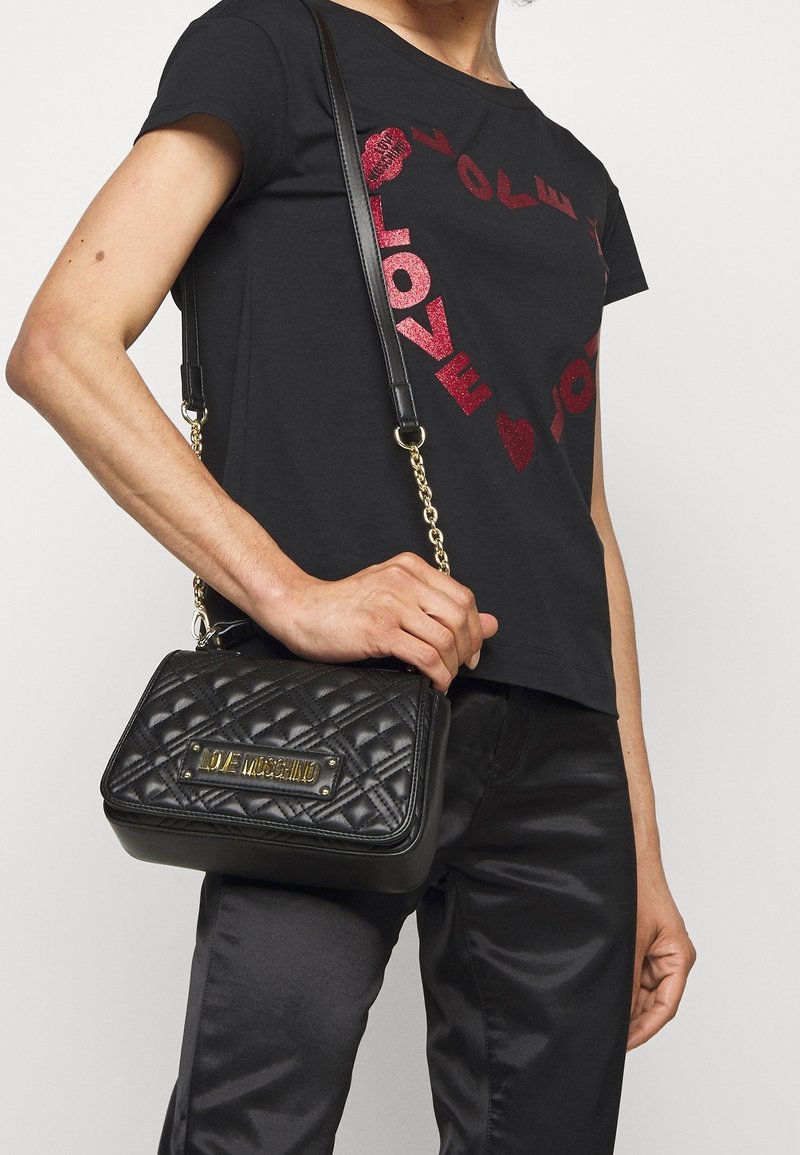 Person bär en svart Love Moschino kviltad crossbody-väska med guldkedja och svart t-shirt med röd glittrande "LOVE"-text och hjärtformad design.