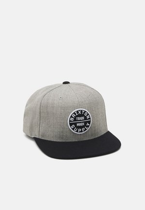 Grå snapback kasket med sort flad skygge og cirkulært mærke med teksten "Brixton Supply Trade Mark" på frontpanelet.