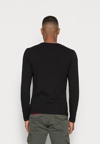 Camisa negra de manga larga con cuello redondo, ajustada, hecha de tela suave, presentada desde la parte posterior sobre un fondo blanco liso.