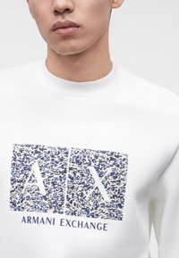 Νέος άνδρας που φοράει λευκό φούτερ Armani Exchange με υφασμένους μπλε και λευκούς μπλοκ που δείχνουν τα γράμματα Α και Χ μπροστά.