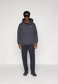 Puffer jacket in dark gray, featuring a hood, two front pockets, elastic cuffs, and a logo on the chest. Paired with matching dark pants.