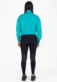 Felpa turchese corta con colletto alto, abbinata a leggings neri con dettagli in rete. Calzature nere completano l'outfit.